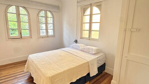 Ein Schlafzimmer mit einem Bett, zwei Fenstern, Holzboden und einer weißen Tür.
