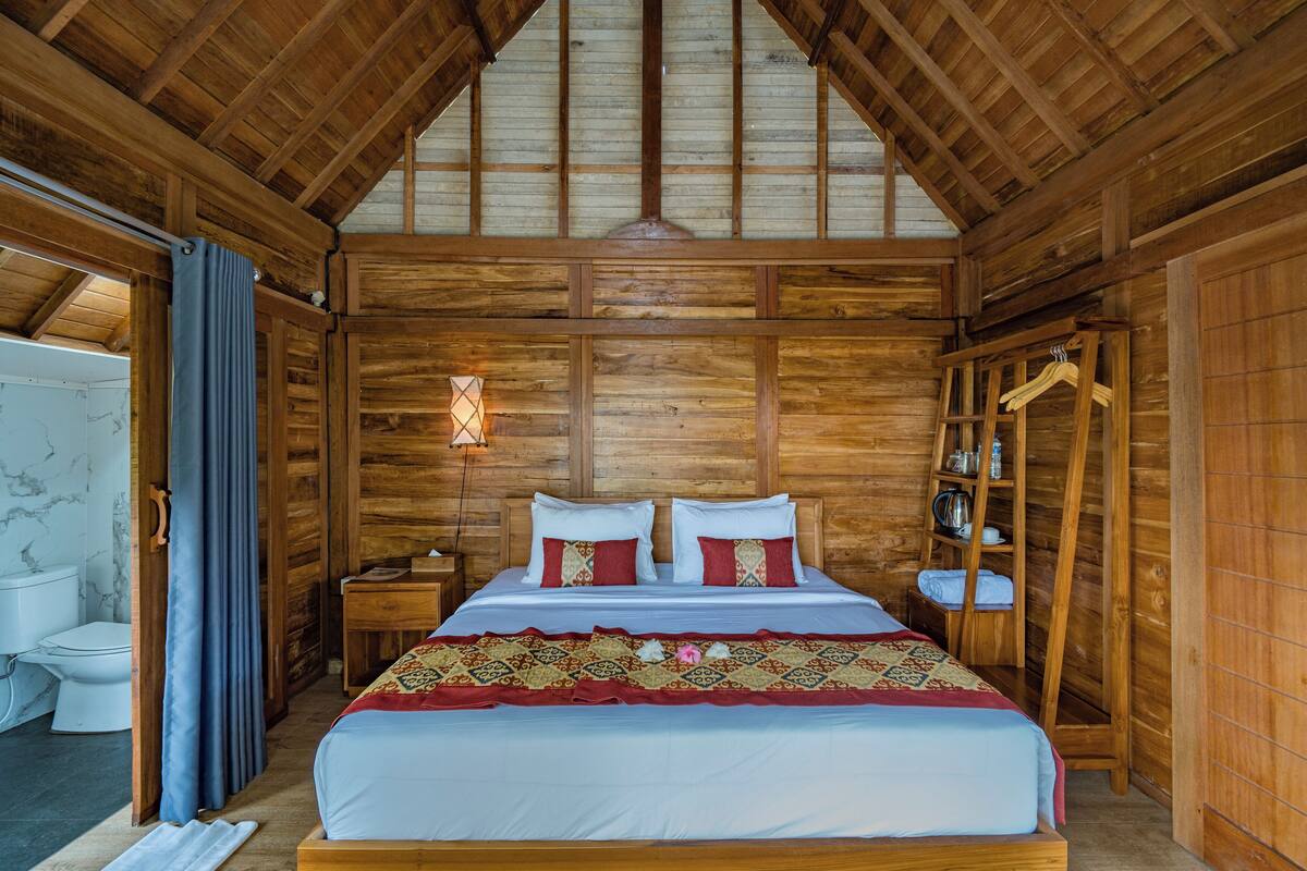 Quarto de cabana de madeira com cama, mesa de cabeceira, prateleira com livros e um cabideiro.