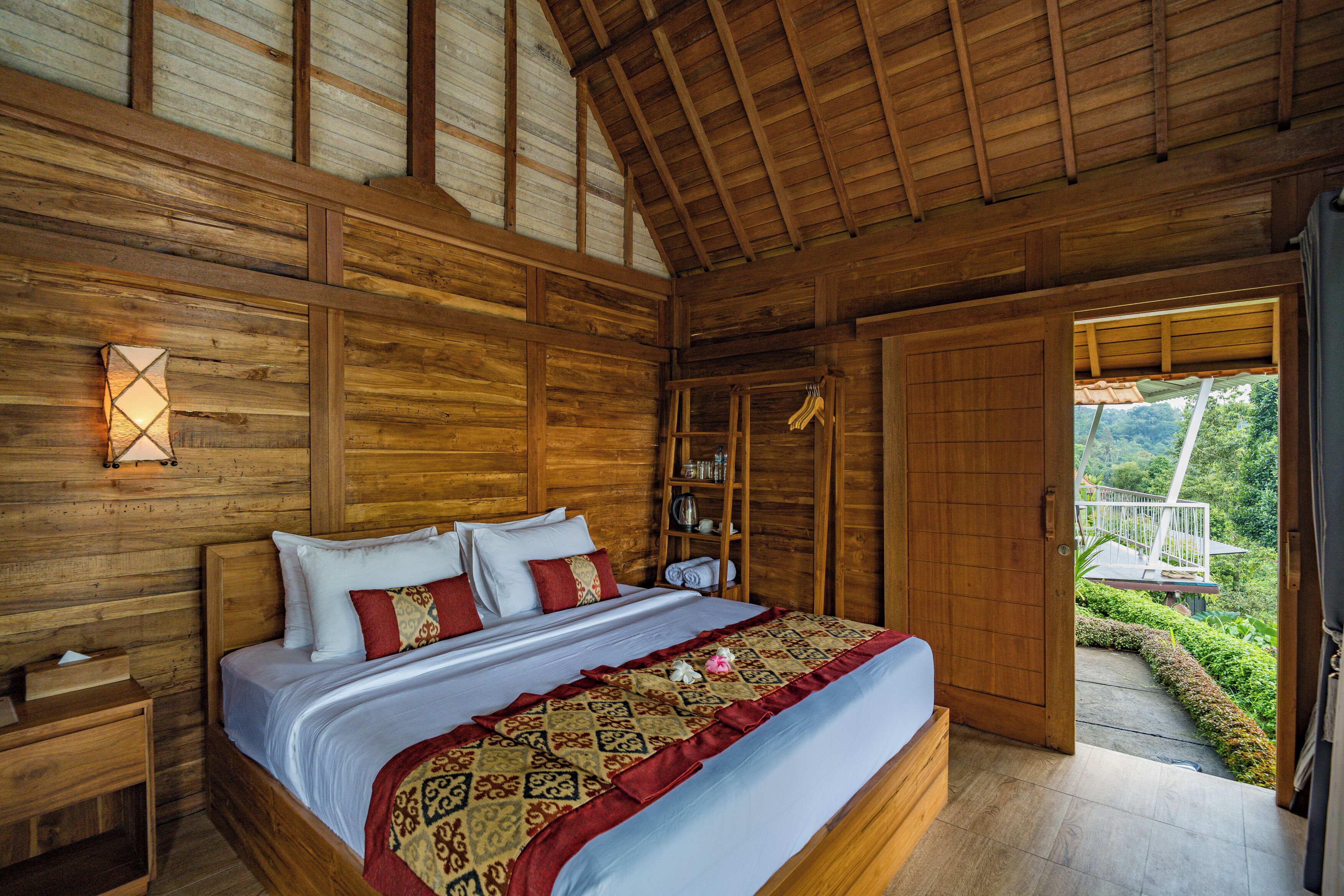 A wooden room with a bed, a nightstand, a shelf with books, and a door leading to an outdoor area.