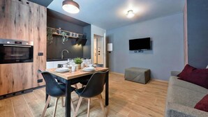 Una cocina moderna con horno integrado, mesa de comedor con sillas, televisor montado en la pared y un sofá gris con cojines rojos.