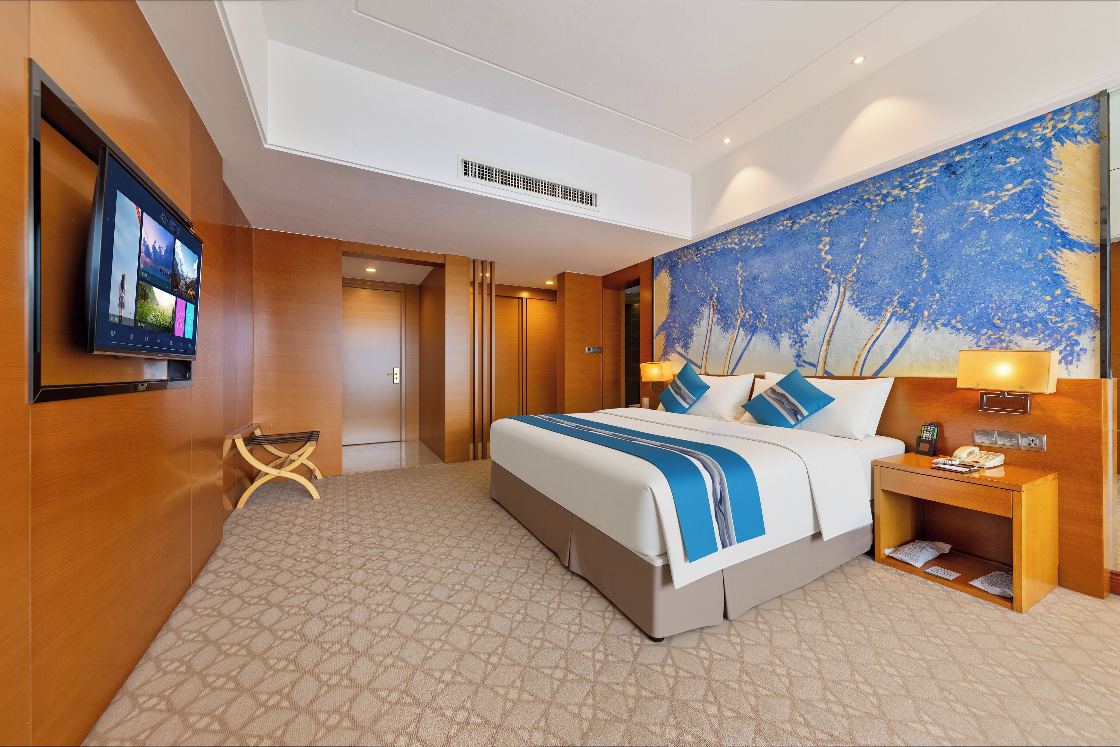 A hotel room with a large bed, a flat-screen TV mounted on the wall, a painting on the wall, and a wooden nightstand with a lamp.