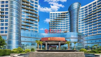 A modern hotel with a prominent glass facade, a central entrance with a red awning, and a landscaped area with a water feature in front.