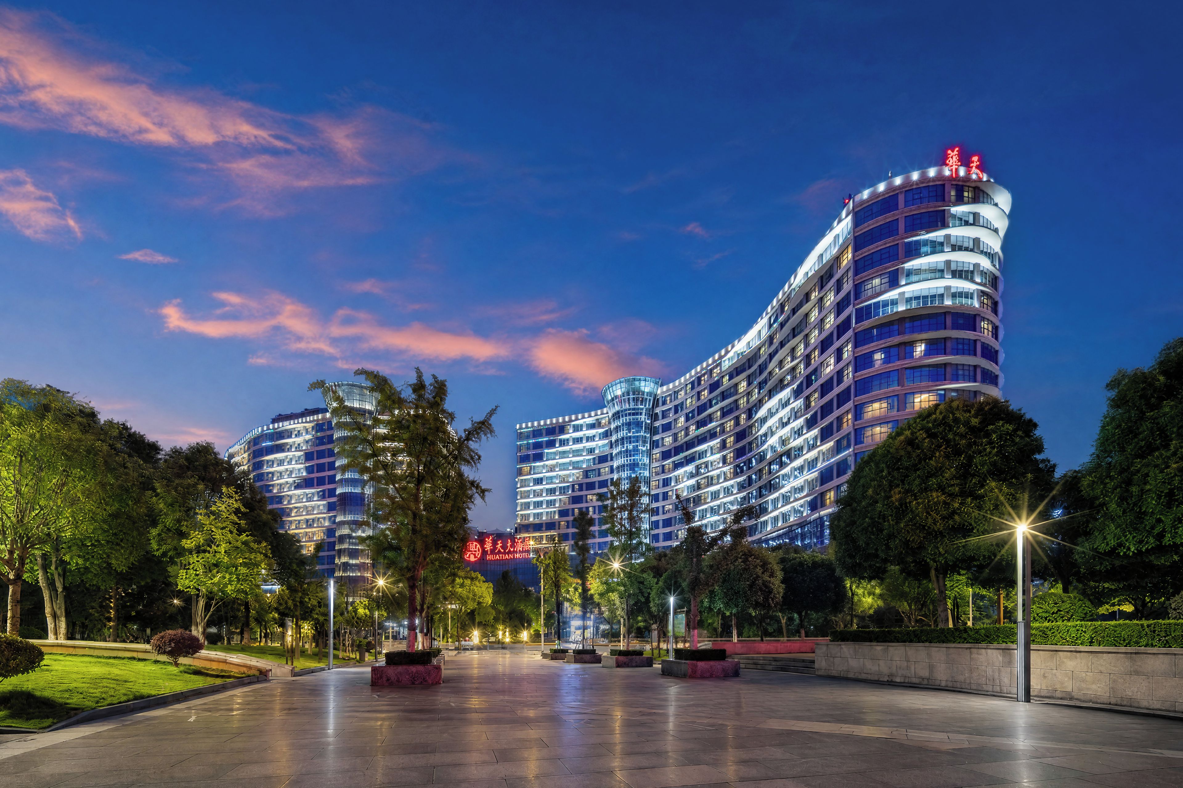 A modern hotel with a curved facade, multiple floors, and a well-lit entrance area with trees and benches.