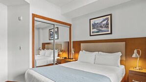 Habitación de hotel con una cama grande, cabecera de madera, mesita de noche con lámpara, espejo y un cuadro en la pared.
