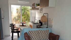 Un salon moderne avec un canapé, un coin repas avec des chaises, une cuisine équipée d’un évier et d’une cuisinière, et une grande fenêtre donnant sur l’extérieur, où se trouvent des plantes.