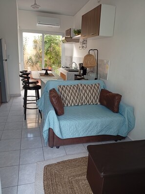 A modern living room with a sofa, a dining area with chairs, a kitchen with a sink and stove, and a large window with plants outside.