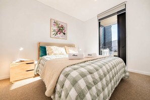 A bedroom with a bed, bedside tables, a nightstand, a window with a view, and a wall-mounted artwork. - Welles St | Christchurch CBD | Sleeps 4 | Balcony (Christchurch)