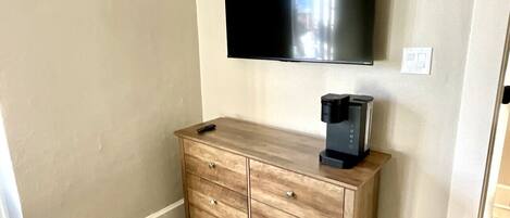 Een kamer met een wandgemonteerde flatscreen-tv, een houten dressoir met lades, en een koffiezetapparaat erop.