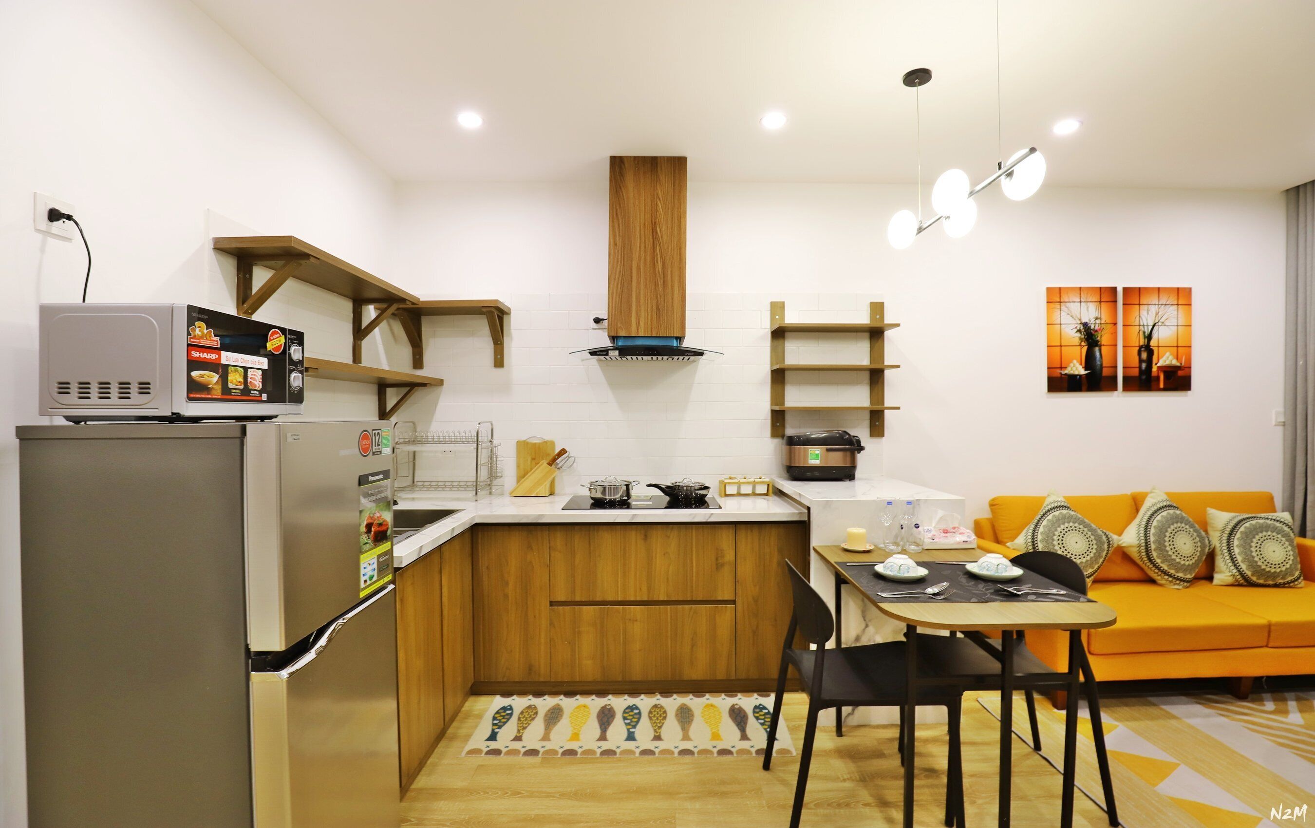 Cozinha moderna com forno embutido, micro-ondas e frigorífico. Há uma mesa de jantar de madeira com cadeiras e um sofá amarelo na sala de estar.
