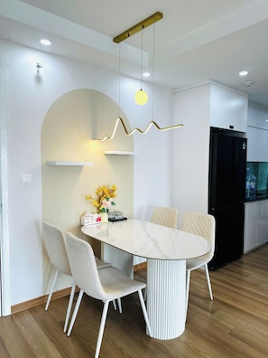 A modern kitchen with a dining area, featuring a marble table, white chairs, a hanging light fixture, and a black refrigerator. - Tami Stay - Vinhomes D'Capitale (Cau Giay)