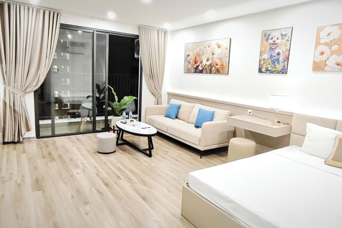 A modern hotel room with a sofa, a round coffee table, a bed, a desk, and a sliding glass door to the balcony.