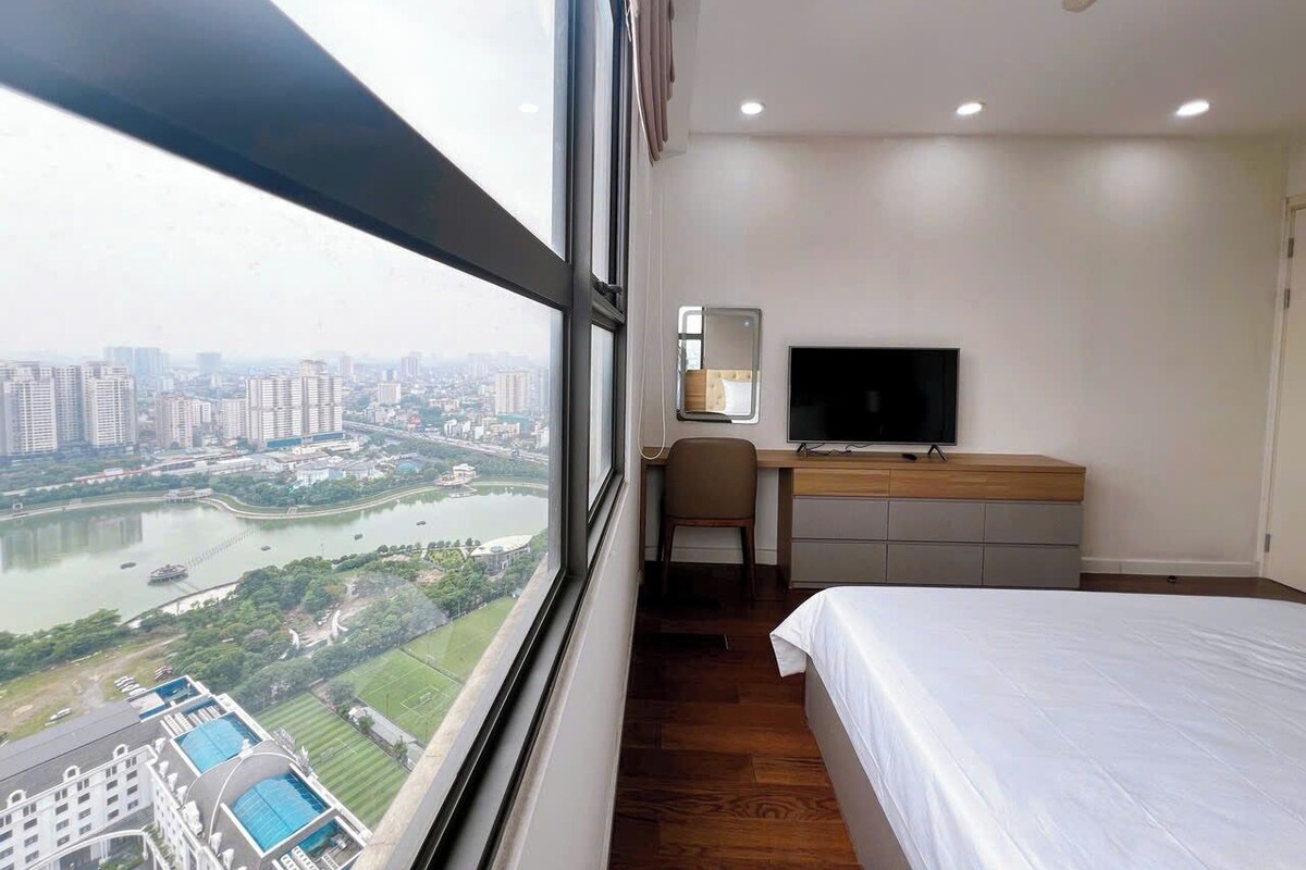 A hotel room with a large window, a bed, a desk with a TV, and a view of a cityscape and river.