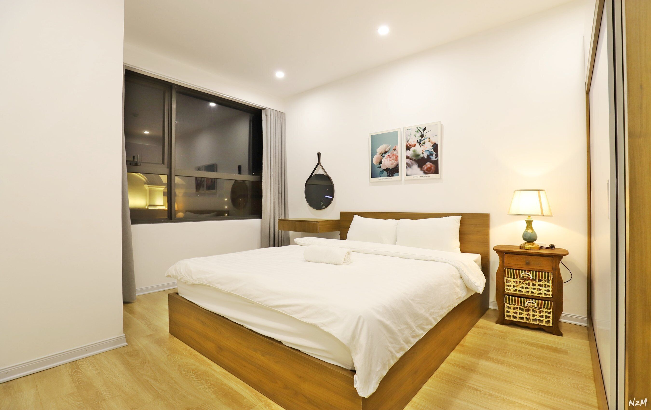 A hotel room with a wooden bed, a bedside table with a lamp, a wall clock, and a window with curtains.