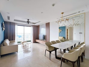 A modern living room with a sofa, a round coffee table, a dining table with chairs, a TV, and a large window with curtains. - Tami Stay - Vinhomes D'Capitale (Cau Giay)
