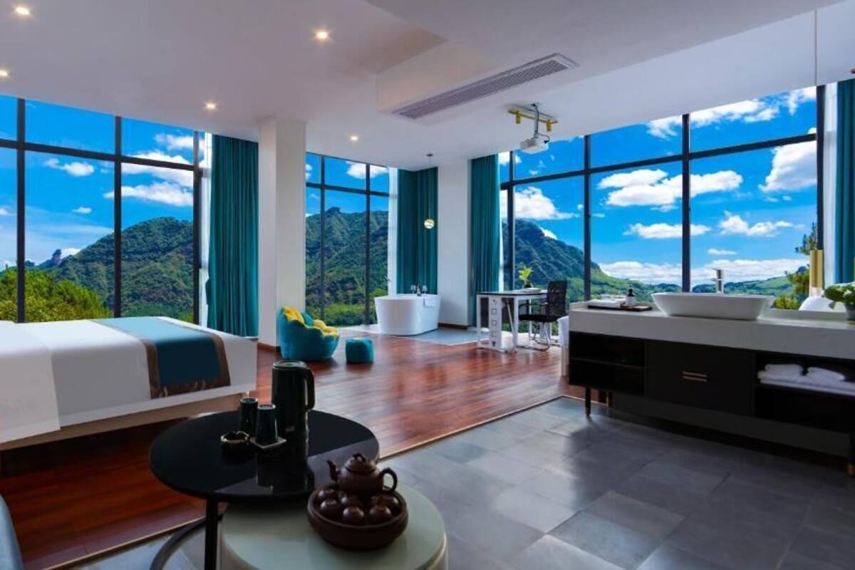 A modern hotel room with a large bed, a seating area, a dining table, and a view of mountains through large windows.