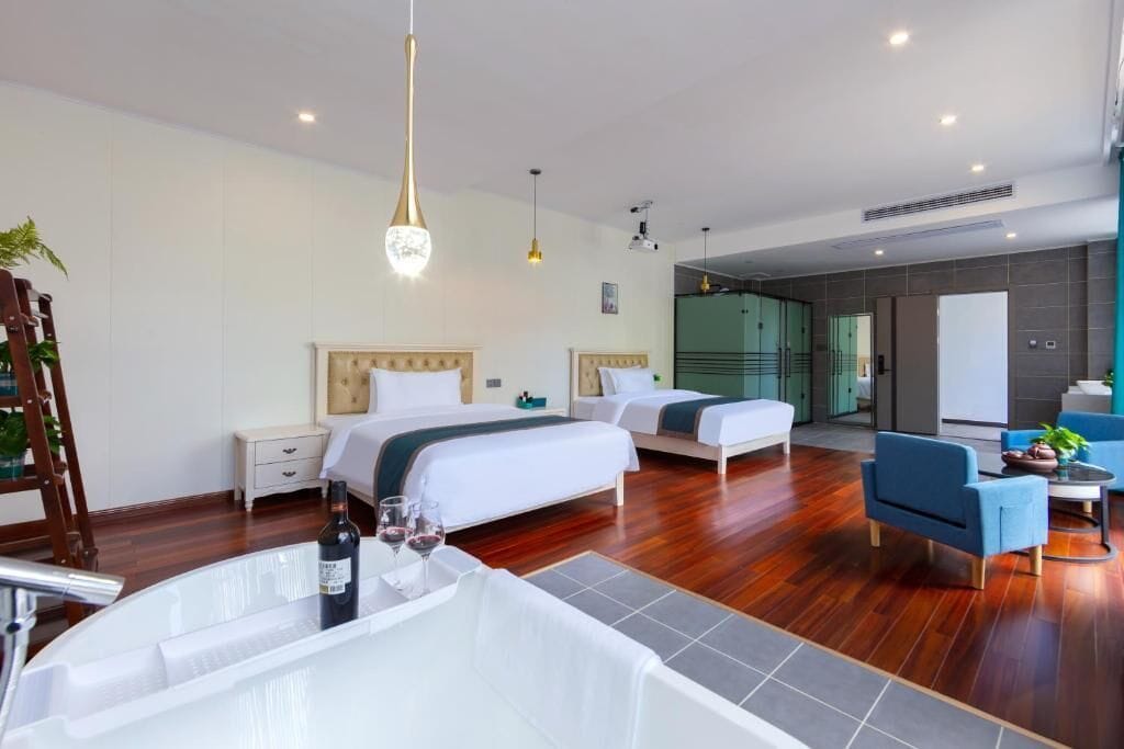 Una habitación de hotel moderna con dos camas, una zona de estar con una silla azul, una mesita pequeña y una bañera grande.