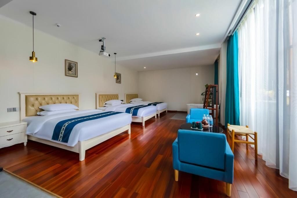 A hotel room with three beds, a blue armchair, a small table, and a large window with curtains.