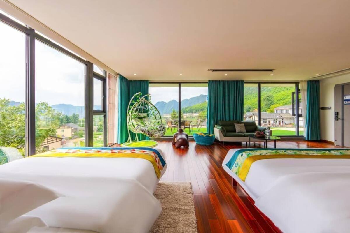 A hotel room with two beds, a large window, a hanging chair, and a view of greenery and mountains.