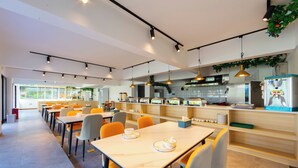 Un espace de salle à manger moderne avec des tables et des chaises, une cuisine en arrière-plan et des luminaires suspendus.