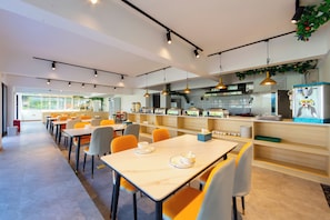 Un comedor moderno con mesas y sillas, una cocina al fondo e iluminación colgante.