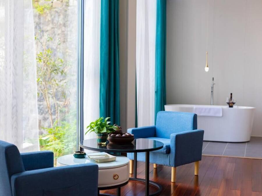Una habitación moderna con una mesa redonda, dos sillones azules, una mesita auxiliar blanca, una planta en maceta y vistas al exterior con vegetación.