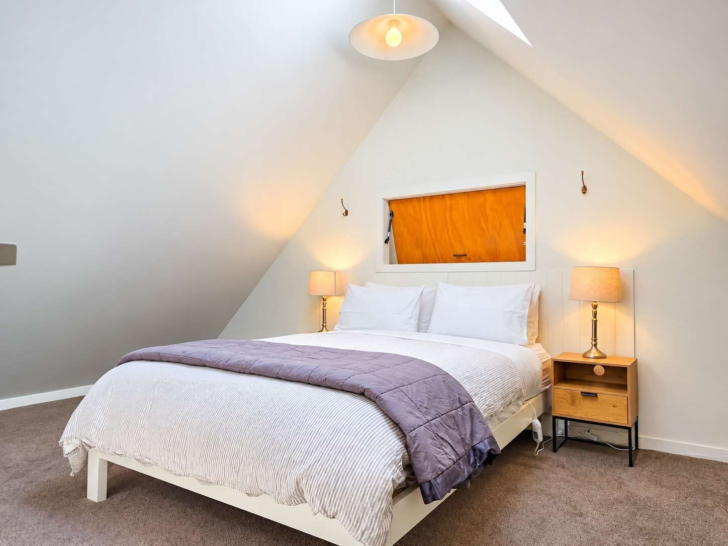 A bedroom with a sloped ceiling, a bed with a white headboard, two bedside tables with lamps, and a wooden door.