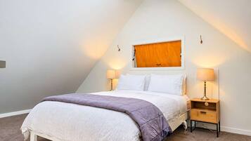 A bedroom with a sloped ceiling, a bed with a white headboard, two bedside tables with lamps, and a wooden door.