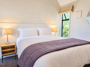 A bedroom with a bed, two bedside lamps, a nightstand, and a window with a view of greenery. - Starfall | Lake Views + Cedar Tub | 2BR (Lake Tekapo)