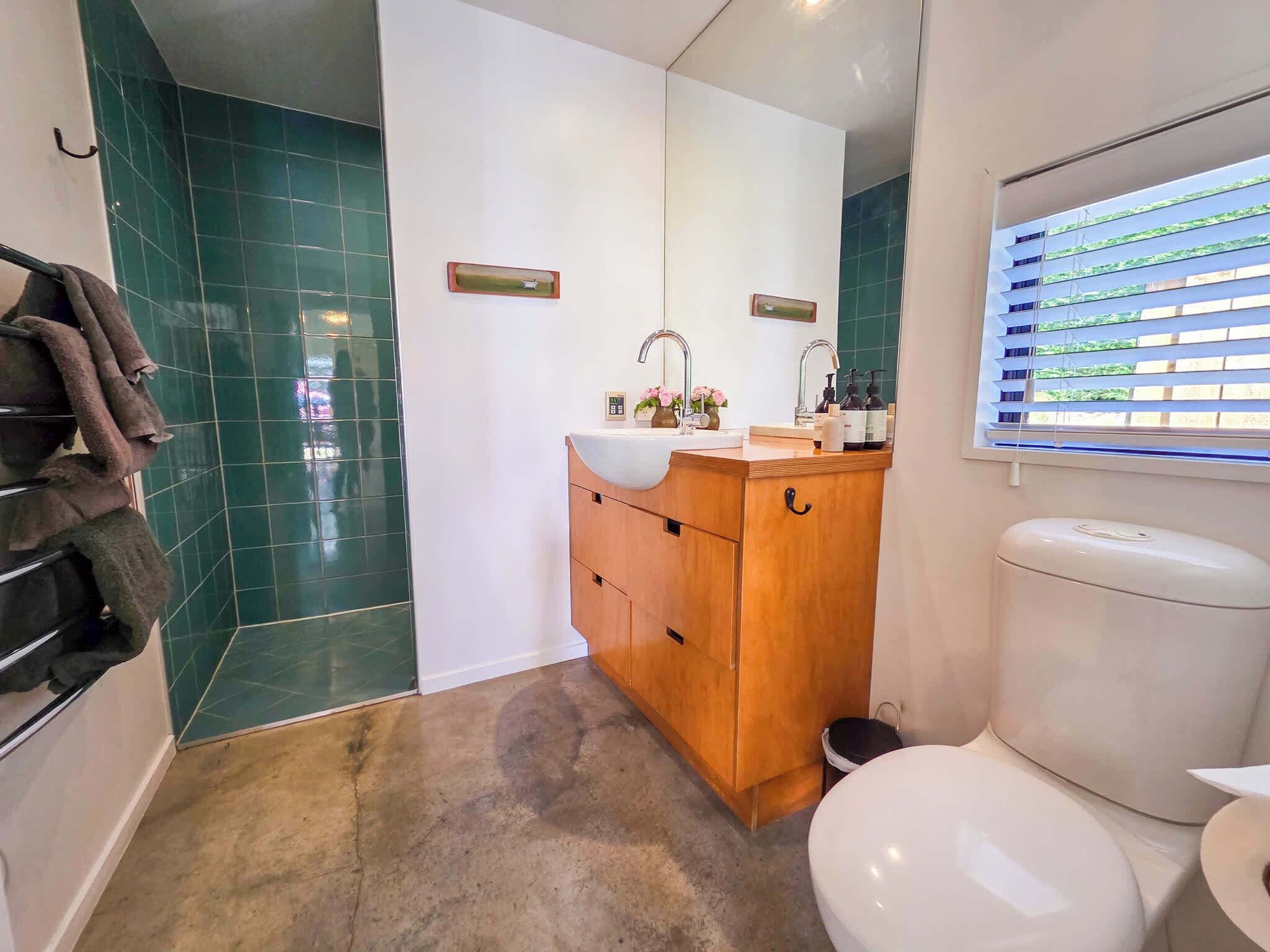 Un baño con inodoro blanco, tocador de madera, paredes con azulejos verdes y una ventana con persianas.