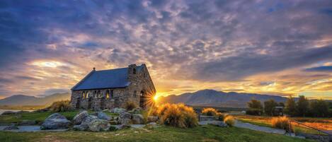 A stone building with a sloped roof, surrounded by grass and rocks, with mountains in the background.