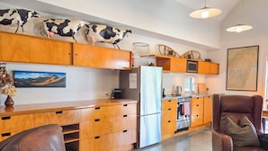 A modern kitchen with wooden cabinets, a refrigerator, a stove, and a microwave. There are two chairs and a table in the living area. - The Black House Horizon Retreat -Private Outdoor Tub (Lake Tekapo)
