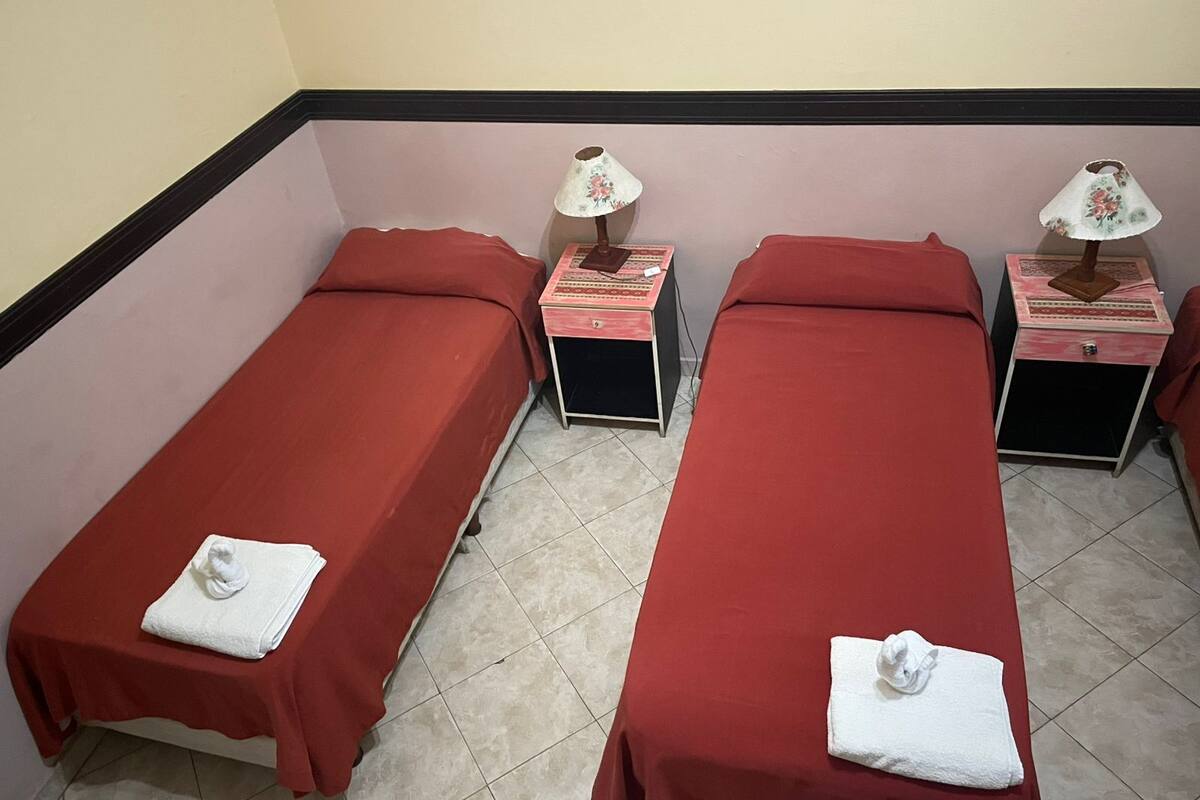 A room with two single beds, each with a red bedspread and a white towel. There are two bedside tables with lamps and a small box on each. The walls are painted in a light color, and the floor is tiled.