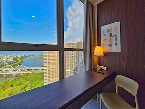 A hotel room with a desk, chair, lamp, and a large window offering a view of a bridge and greenery. - By The Sky By SanLiving - 3BR - PIK (Jakarta)