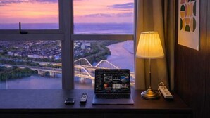 Une chambre d’hôtel avec une grande fenêtre, un bureau avec un ordinateur portable, une lampe, une télécommande et un téléphone.
