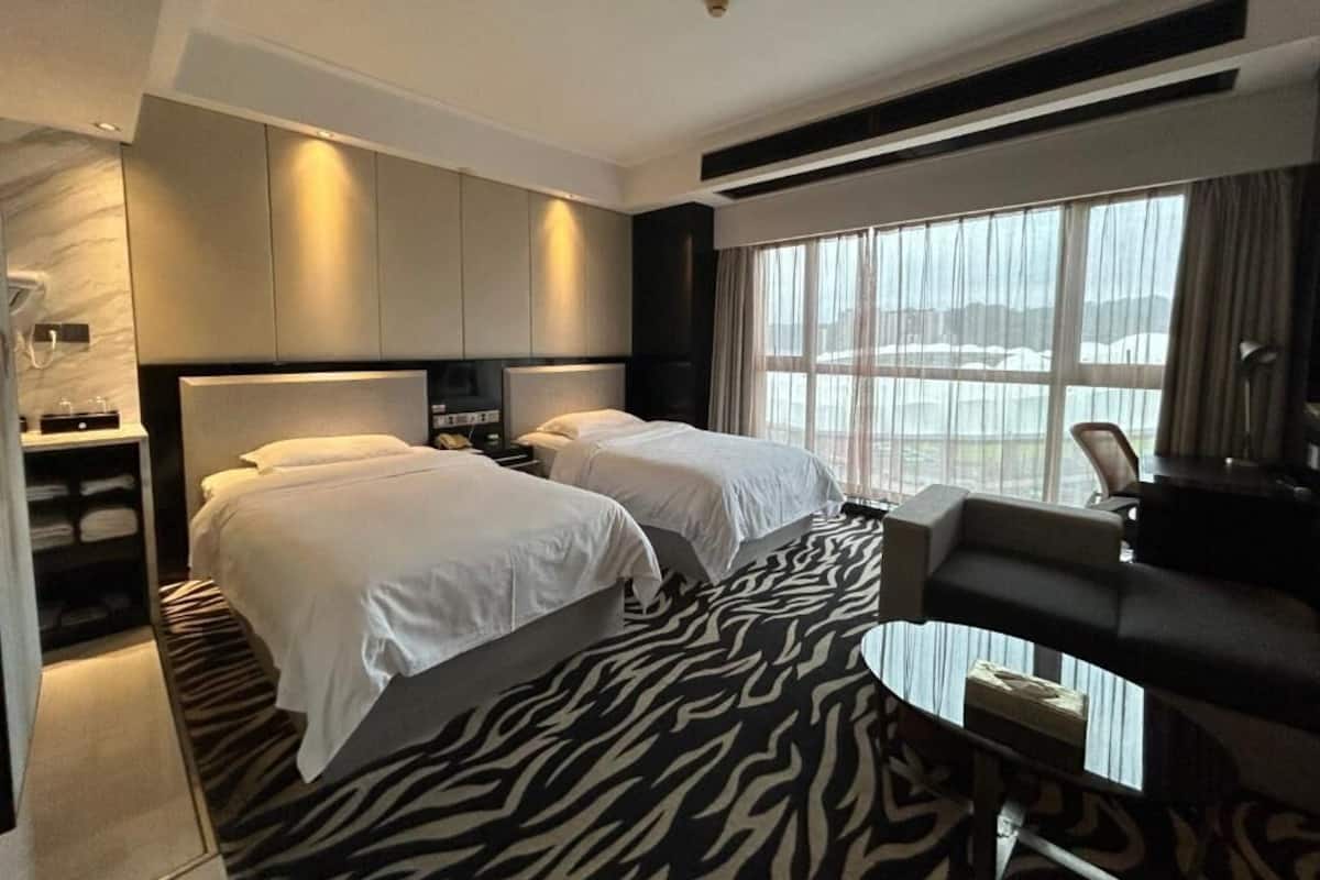 Habitación de hotel con dos camas, un escritorio, una silla, un ventanal grande con cortinas y una alfombra estampada con motivos de cebra.