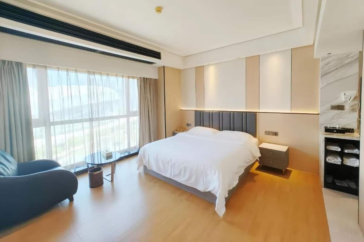 Habitación de hotel moderna con una cama grande, una mesita de noche, una silla y vistas al exterior a través de amplias ventanas.