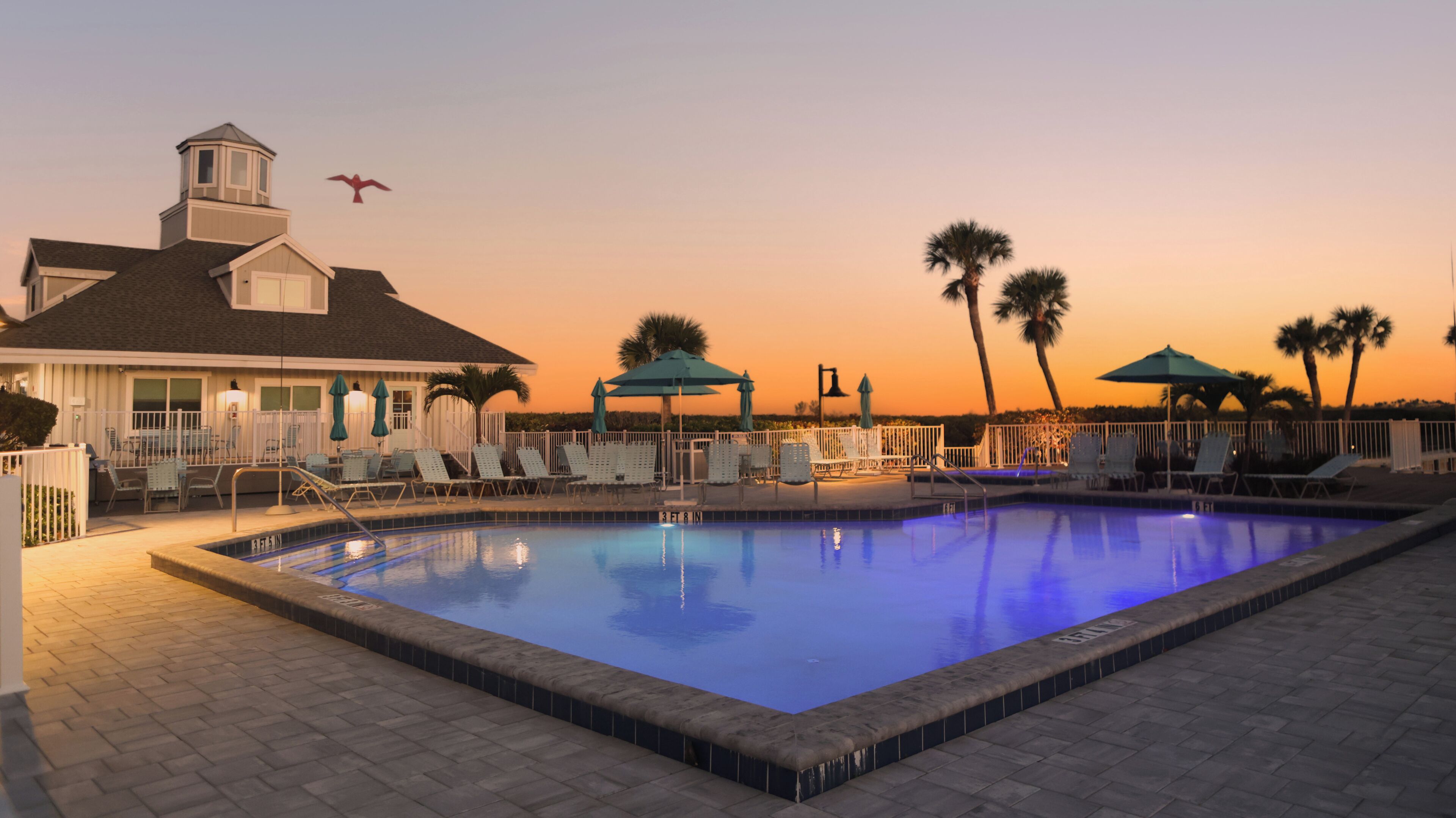 Ein Swimmingpool mit Liegestühlen und Sonnenschirmen, ein Gebäude mit einem Turm sowie Palmen vor einem Sonnenuntergangshimmel.