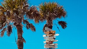 A beach with palm trees, a wooden signpost with destination names, and a picnic table. - Rare BOCA GRANDE 3BR/3BA WATERFRONT - Gulf Sunsets & Harbor Sunrises - Boat Slip (Boca Grande)