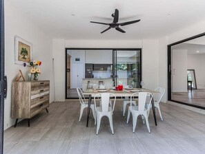 Ein Essbereich mit einem Tisch, gedeckt für vier Personen, weißen Stühlen, einem Deckenventilator und Blick in die Küche durch Glastüren.