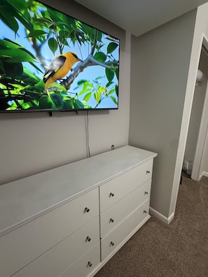 Ein an der Wand befestigter Fernseher, vor dem ein weißes Schminktischchen und ein Teppichboden zu sehen sind.
