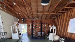 Een ruime kamer met een plafondventilator, een zwarte trainingsmachine, een witte luchtreiniger en een zwarte bank met gestreepte kussens.