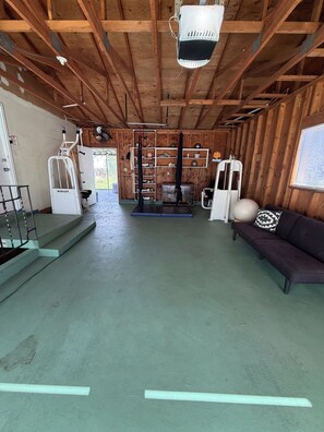 A spacious room with a ceiling fan, a black exercise machine, a white air purifier, and a black sofa with patterned cushions. - The Garden Billiard Club (Orem)