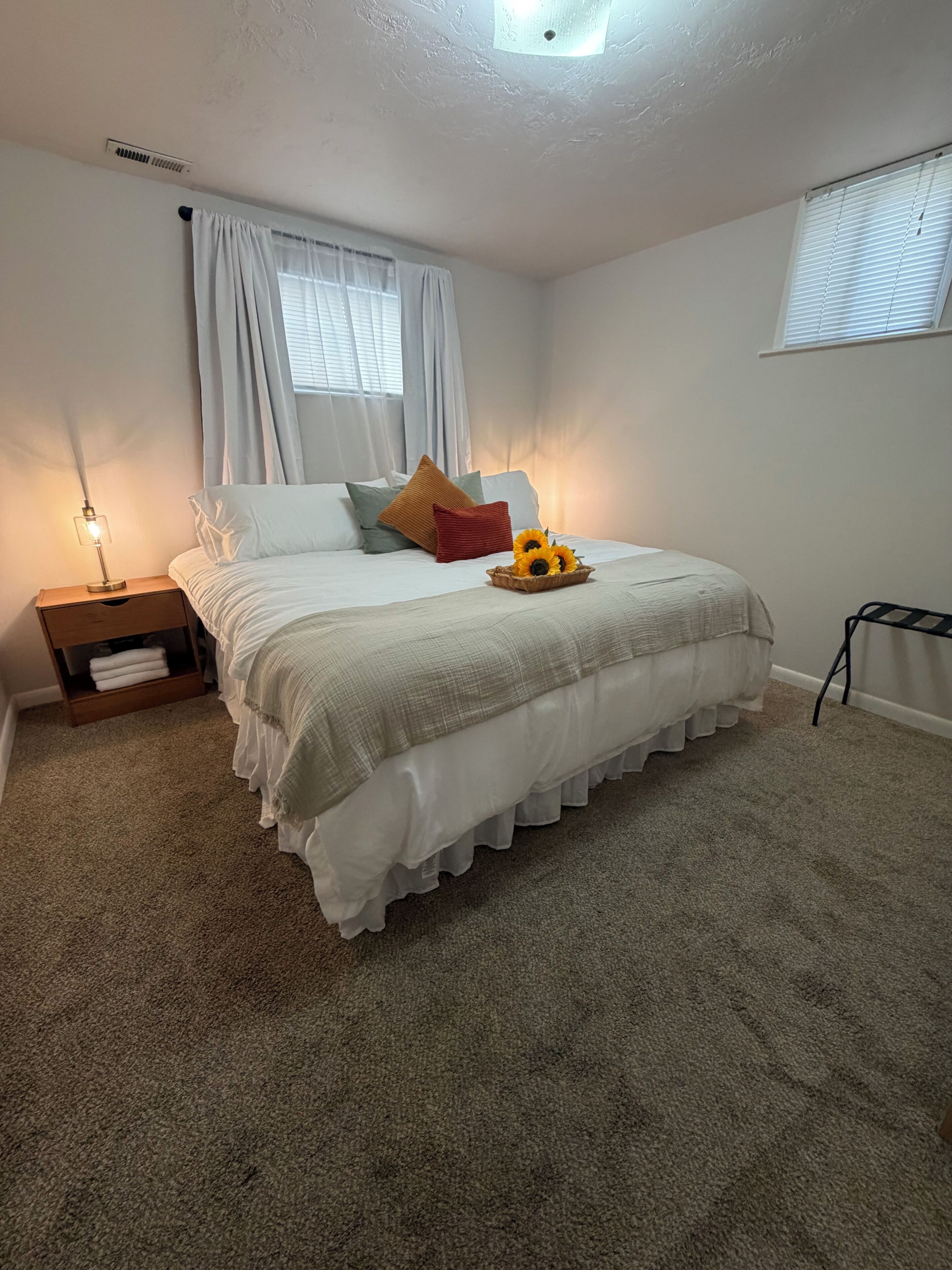 Un dormitorio con cama, mesita de noche, ventana con cortinas y una pequeña mesa con lámpara.