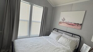 Un dormitorio con cama, cortinas, una ventana, una mesita de noche y un cuadro colgado en la pared.