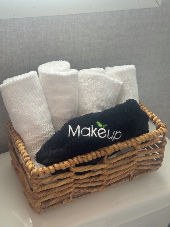 Una cesta tejida con toallas blancas y una toalla negra con el bordado 'Makeup'.