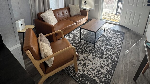 Una sala de estar moderna con un sofá de cuero marrón, una silla de madera, una mesita de centro y un tapete estampado.
