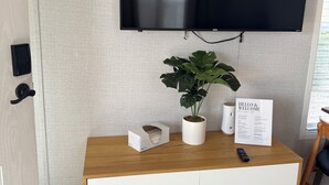Una habitación moderna con una televisión montada, un mueble de madera con cajones, una planta en maceta, un cartel de bienvenida y un control remoto sobre el mueble.