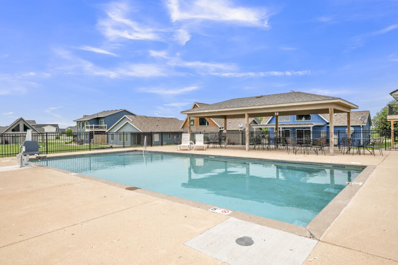 A rectangular swimming pool with a tiled deck, surrounded by a fence and a covered patio area with chairs.