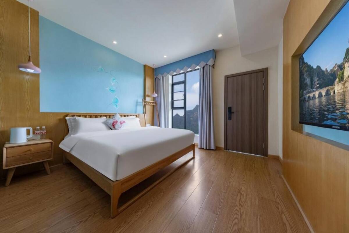 Un dormitorio con una cama grande, suelo de madera, una mesita de noche con lámpara, una ventana con cortinas y un cuadro en la pared.