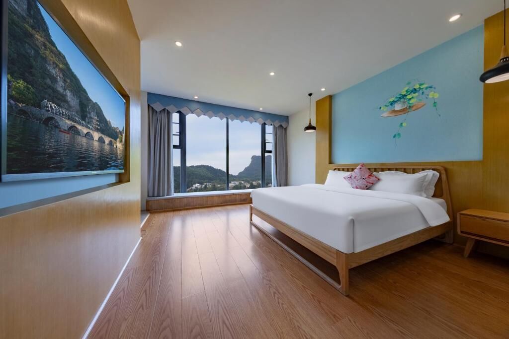 A bedroom with a large window, a bed, a nightstand, and a painting on the wall.